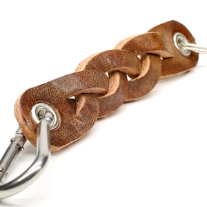 Może przedstawiać: Pleciony brelok do kluczy ze skóry w kolorze brązowym ze srebrnym karabińczykiem i kółkiem na klucze. Skóra ma ciepły, naturalny brązowy kolor, a metalowe okucia są błyszczące. Brelok jest przeznaczony do przechowywania kluczy lub innych drobnych przedmiotów.