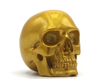 Pode incluir: Uma figurinha de caveira pintada de ouro. A caveira é detalhada com órbitas oculares, um nariz e dentes.