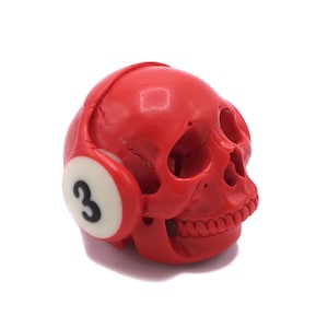 May include: A red skull ornament with headphones. The headphones have white earcups with the number 3 in black. The skull has detailed features and is set against a white background.