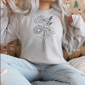 May include: A gray sweatshirt with a black line drawing of a bouquet of flowers. The flowers are stylized and include a daisy, a smaller flower with five petals, and a flower with a center circle. The text "Trust the timing" is written vertically within the bouquet.
