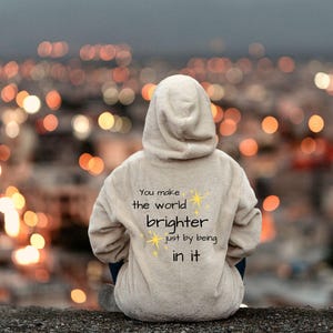May include: A person wearing a light beige hoodie with the text "You make the world brighter just by being in it" in black and yellow. The person is sitting with their back to the camera, overlooking a blurred cityscape with bokeh lights.