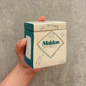 以下が含まれることがあります： 手描きのセラミックソルトボックス、蓋付き。ボックスは白で、ティールアクセントがあり、「THE ORIGINAL」と「Maldon SEA SALT！」の文字がひし形に配置されています。ボックスには「250g」と記載されています。