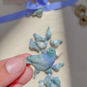 Op de afbeelding: Een verzameling kleine, handgeschilderde keramische vogels in tinten blauw en groen. De vogels zijn in een cluster gerangschikt, met één vogel in een hand.