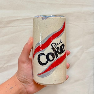 Op de afbeelding: Een keramische beker in de vorm van een Diet Coke-blikje. De gebroken witte beker heeft het Diet Coke-logo in zwart en rood, met grijze accenten. De beker wordt door een hand vastgehouden.