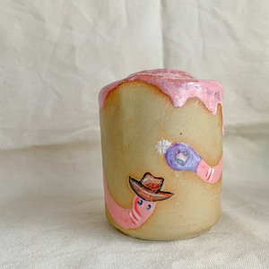 May include: A ceramic mug with a pink glaze and two painted pink worms. One worm is wearing a cowboy hat and the other is wearing a purple hat.