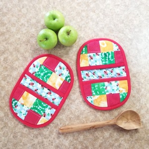 May include: Two red quilted pot holders with a patchwork design featuring green, yellow, and blue fabric with a ladybug print. The pot holders are on a light brown countertop with three green apples.