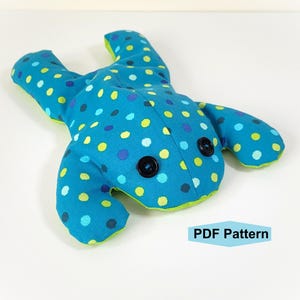 Puede incluir: Un peluche con forma de rana de tela turquesa con un estampado de lunares en azul, verde y amarillo. La rana tiene dos ojos de botón negros y ribetes verdes. El texto "PDF Pattern" está en un recuadro azul.