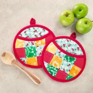 May include: Two red and white quilted pot holders with a pocket on each. The pot holders have a patchwork design with green, yellow, and blue squares. The pot holders are on a light gray surface.