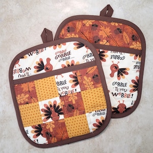 May include: Two brown quilted oven mitts with a patchwork design featuring turkeys, leaves, and polka dots. The mitts have a pocket on the back and the text "Gobble Til You Wobble!" is printed on the front.