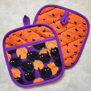 Może przedstawiać: Para rękawic kuchennych w kolorze fioletowym i pomarańczowym z motywem Halloween. Jedna rękawica ma wzór z czarnym kotem i dynią, a druga rękawica ma wzór z czarnym kotem i pomarańczową dynią.
