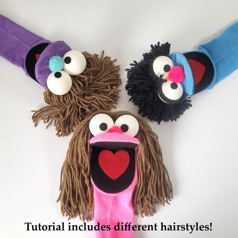 May include: Three sock puppets with yarn hair, large white ping pong ball eyes, and felt mouths. One puppet has a purple sock, one blue, and one pink. The pink puppet features a red heart inside its mouth. Text reads: Tutorial includes different hairstyles!