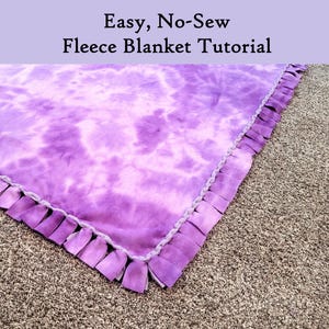 Puede incluir: Una manta de forro polar teñida con efecto tie-dye en tonos morados, con un borde de flecos anudados. El texto "Easy, No-Sew Fleece Blanket Tutorial" aparece en la parte superior. La manta se muestra sobre una superficie texturizada de color neutro.