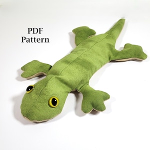 May include: A plush green lizard toy with yellow eyes and a light green belly. The toy is lying on a white surface. The text "PDF Pattern" is in the upper left corner. The lizard has four legs and a long tail.