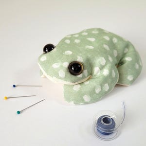 May include: A frog-shaped pin cushion made from light green fabric with white spots. It has large black eyes. Sewing pins with blue, yellow, and green heads are nearby, along with a spool of blue thread.