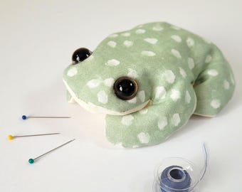 May include: A handmade frog-shaped pin cushion in light green fabric with white polka dots. The frog has large black eyes and is surrounded by sewing pins with blue, yellow, and green heads, and a spool of blue thread.