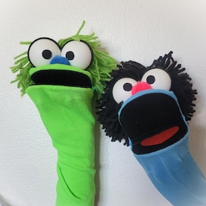 May include: Two sock puppets with yarn hair and large white eyes. The left puppet is bright green with a blue nose and black mouth. The right puppet is light blue with black hair, a pink nose, and a black mouth with a red tongue.