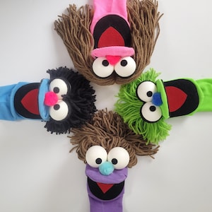 May include: Four sock puppets arranged in a cross pattern. Each puppet features large white spherical eyes, yarn hair, a fabric mouth with a red interior, and a small pom-pom nose. The puppets are pink, blue, green, and purple.