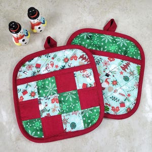 May include: Two red and green Christmas-themed pot holders with a quilted design. One pot holder has a patchwork design with red squares and green and blue squares with snowflakes and Christmas trees. The other pot holder has a green and blue design with snowflakes, Christmas trees, and snowmen.