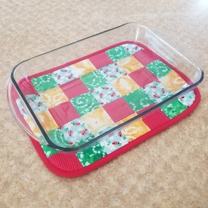 May include: A glass baking dish sits on a red and white quilted hot pad with a patchwork design featuring ladybugs and floral patterns.