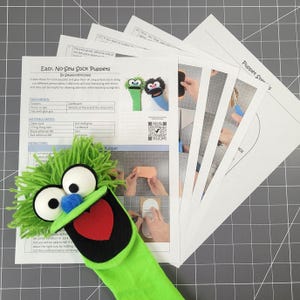 May include: A green sock puppet with large white eyes, a blue nose, and a red heart-shaped tongue, accompanied by a printed instructional guide titled Easy, No-Sew Sock Puppets by SewKnotFinished, featuring step-by-step photos and text.