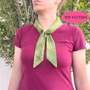 May include: A solid olive green neck scarf tied in a simple knot at the center of the neck. The scarf features pointed ends and a smooth fabric texture. A pink oval graphic in the upper right corner contains the black text PDF PATTERN.