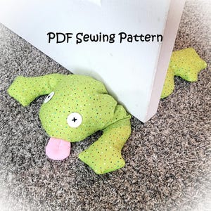 Op de afbeelding: Een groene, kikker-vormige deurstopper met een roze tong, witte ogen en een zwarte kruisknop. De deurstopper is gemaakt van stof met een stippenpatroon. De tekst "PDF Sewing Pattern" staat erboven.