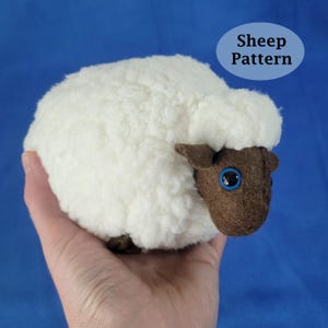May include: A plush sheep toy with a fluffy white body and a brown face and ears. The sheep has large blue eyes and is held in a hand. The text "Sheep Pattern" is in a blue oval.