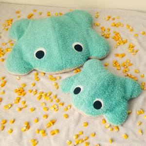 Può includere: Due giocattoli di peluche morbidi, di colore blu turchese, a forma di creature marine. La creatura più grande ha la forma di una manta con due grandi occhi neri. La creatura più piccola ha la forma di una stella marina con due grandi occhi neri.