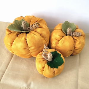 Peut inclure: Trois citrouilles en peluche jaunes de tailles différentes. Chaque citrouille a une feuille en feutre vert et une tige torsadée en forme de corde. Les citrouilles sont disposées sur une surface brun clair, créant un affichage décoratif sur le thème de l'automne.