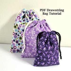 May include: Three drawstring bags with floral patterns in shades of purple, white, and blue. The bags have white or black drawstrings and are arranged in a row. The text "PDF Drawstring Bag Tutorial" is visible.