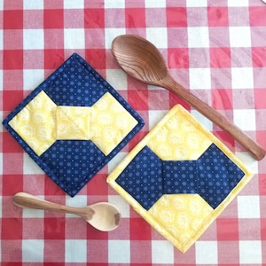 May include: Two yellow and blue quilted pot holders with a bow design. The pot holders are on a red and white checkered tablecloth with a wooden spoon.