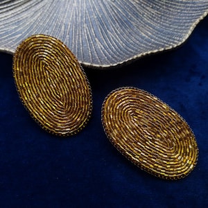 May include: Two gold oval-shaped earrings made of small, shiny beads. The earrings are on a blue background.