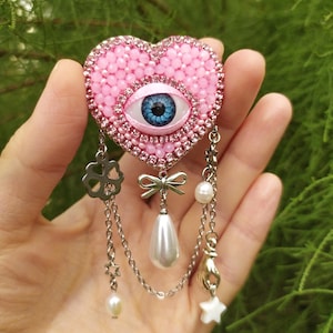 May include: A pink heart-shaped brooch with a blue eye in the center, surrounded by pink beads and rhinestones. Silver chains dangle from the brooch, adorned with charms, pearls, and a bow. A unique and eye-catching accessory.