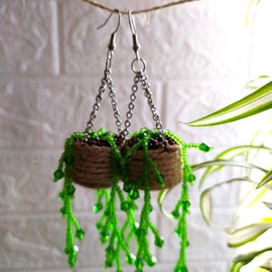 Puede incluir: Un par de pendientes colgantes con una pequeña cesta tejida marrón con plantas de cuentas verdes que cuelgan de ella. Los pendientes están suspendidos de cadenas plateadas.