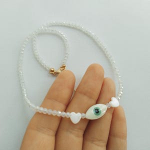 Puede incluir: Un delicado collar de cuentas blancas con un colgante central en forma de ojo con un iris azul y dos acentos blancos en forma de corazón. El collar tiene un cierre dorado.