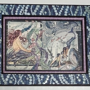 May include: A framed illustration of two mermaids in an underwater scene. The mermaids are surrounded by seaweed, fish, and seashells. The frame is decorated with a blue and white pattern.