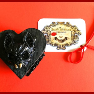 May include: A black heart-shaped box with a bat skull design, featuring gold-colored teeth. A white tag with the text "Amira di Transilvania" and a red ribbon are also present. The box is on a red surface.