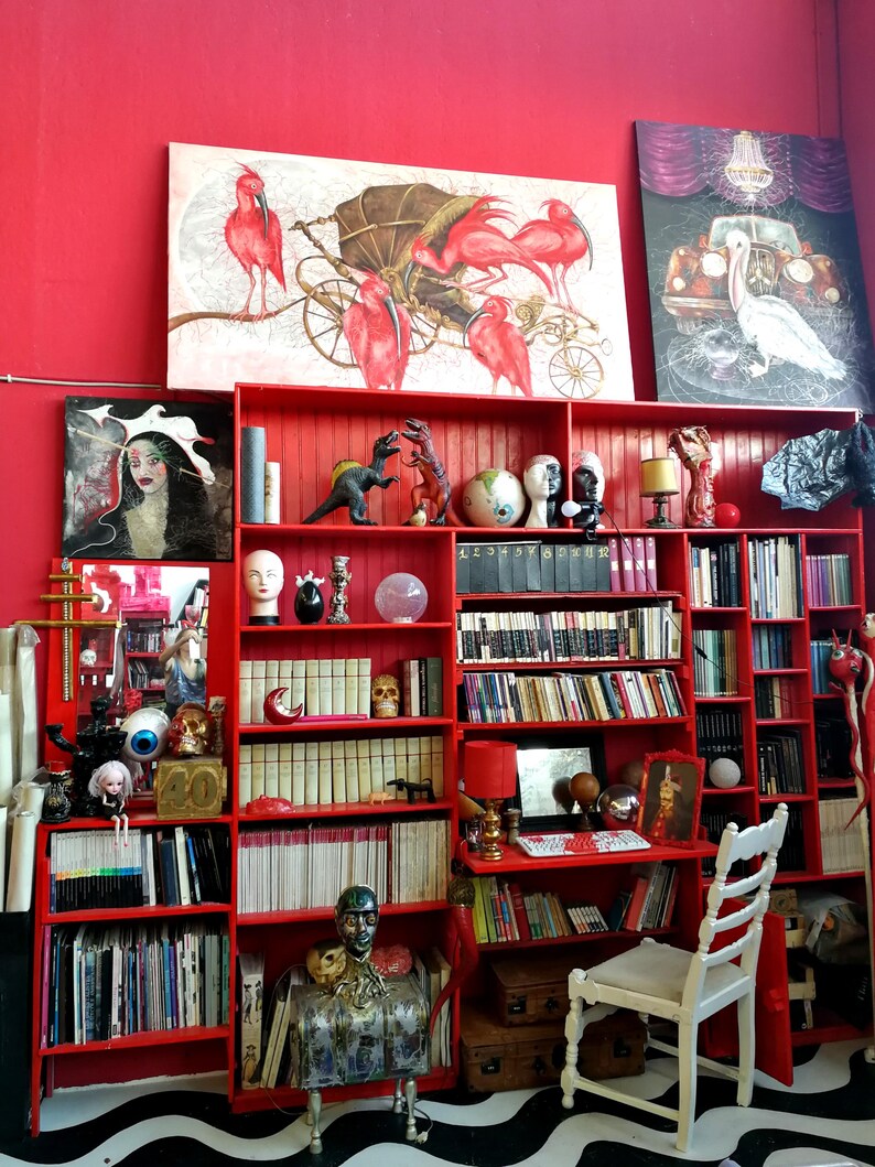 May include: A red bookshelf filled with books and decorative items, including sculptures, globes, and a skull. Artwork featuring birds and a car hangs on the red wall. A white chair sits in front of the bookshelf.