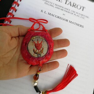 May include: A red, handmade pouch with a decorative tassel and bead accents. The pouch features a circular image with a heart and swords design. The text "Swords" is visible. The background includes a book titled "The Tarot" by S. L. Macgregor Mathers.
