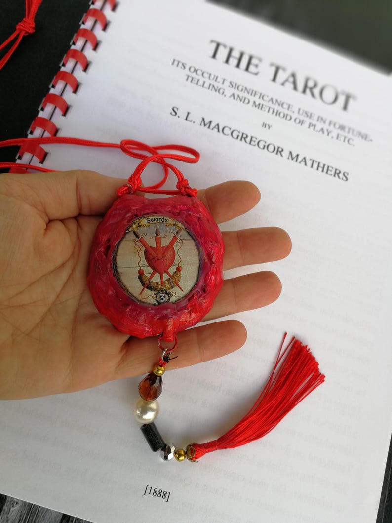 May include: A red, handmade pouch with a decorative tassel and bead accents. The pouch features a circular image with a heart and swords design. The text "Swords" is visible. The background includes a book titled "The Tarot" by S. L. Macgregor Mathers.