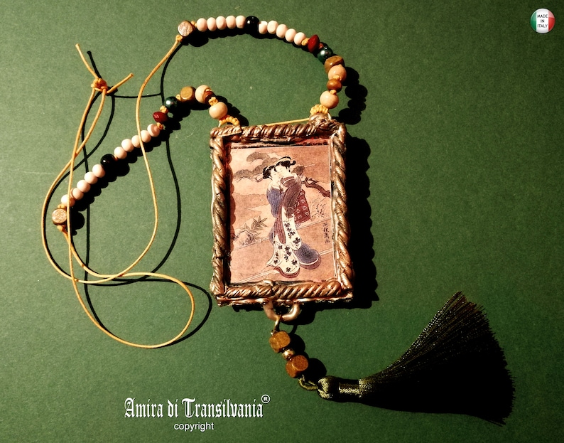 May include: A gold-toned pendant necklace with a framed image of a woman in a traditional Japanese kimono. The necklace features a variety of beads, including white, brown, and green. The pendant has a tassel made of green fabric.