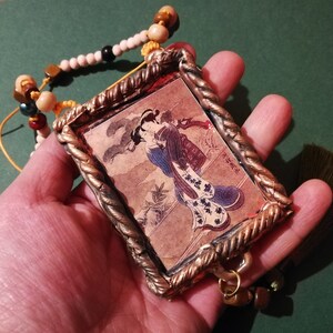 May include: A gold-toned pendant with a framed image of a woman in a blue and white kimono. The pendant is attached to a beaded necklace with brown, white, and yellow beads.