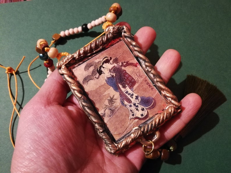 May include: A gold-toned pendant with a framed image of a woman in a blue and white kimono. The pendant is attached to a beaded necklace with brown, white, and yellow beads.