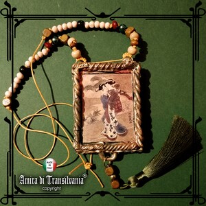May include: A necklace with a silver frame pendant featuring a Japanese woodblock print of a woman in a kimono. The necklace is made with white, brown, and green beads and has a green tassel.