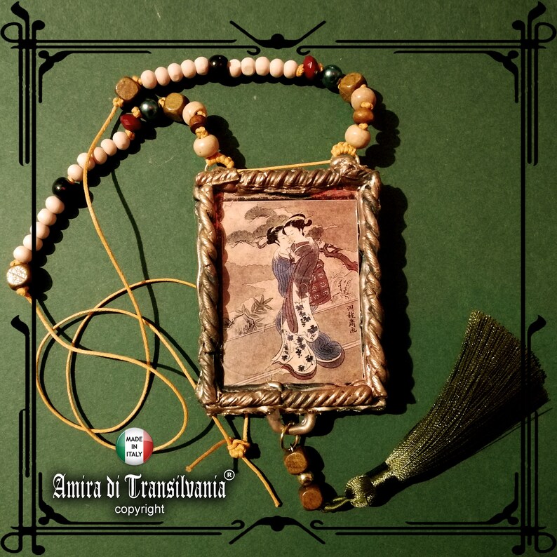 May include: A necklace with a silver frame pendant featuring a Japanese woodblock print of a woman in a kimono. The necklace is made with white, brown, and green beads and has a green tassel.