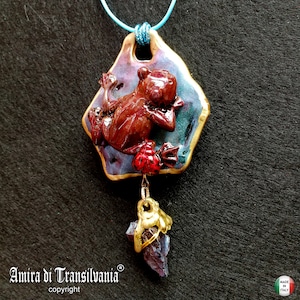 Peut inclure: Un collier avec un pendentif en céramique représentant une grenouille rouge et brune sur un fond violet et bleu. Le pendentif est attaché à une chaîne dorée avec un charme en cristal violet.
