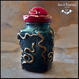 May include: A decorative, hand-painted jar with a dark teal base and gold snake detailing. The jar has a red lid with gold accents. The jar is labeled "Amira di Transilvania" and "Made in Italy."
