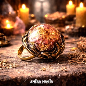 May include: A handcrafted ring with a large, glittery, orb-shaped stone in shades of gold, pink, and red. The ring has an ornate, gold-colored setting. The ring is displayed on a wooden surface with a blurred background of candles and foliage. The text "AMIRA MAGIA" is visible.