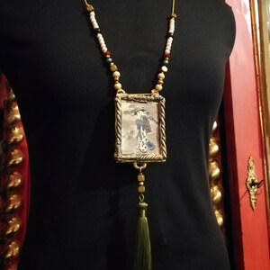 May include: A gold necklace with a small framed picture of a woman in a kimono. The necklace has white, red, and green beads and a green tassel.