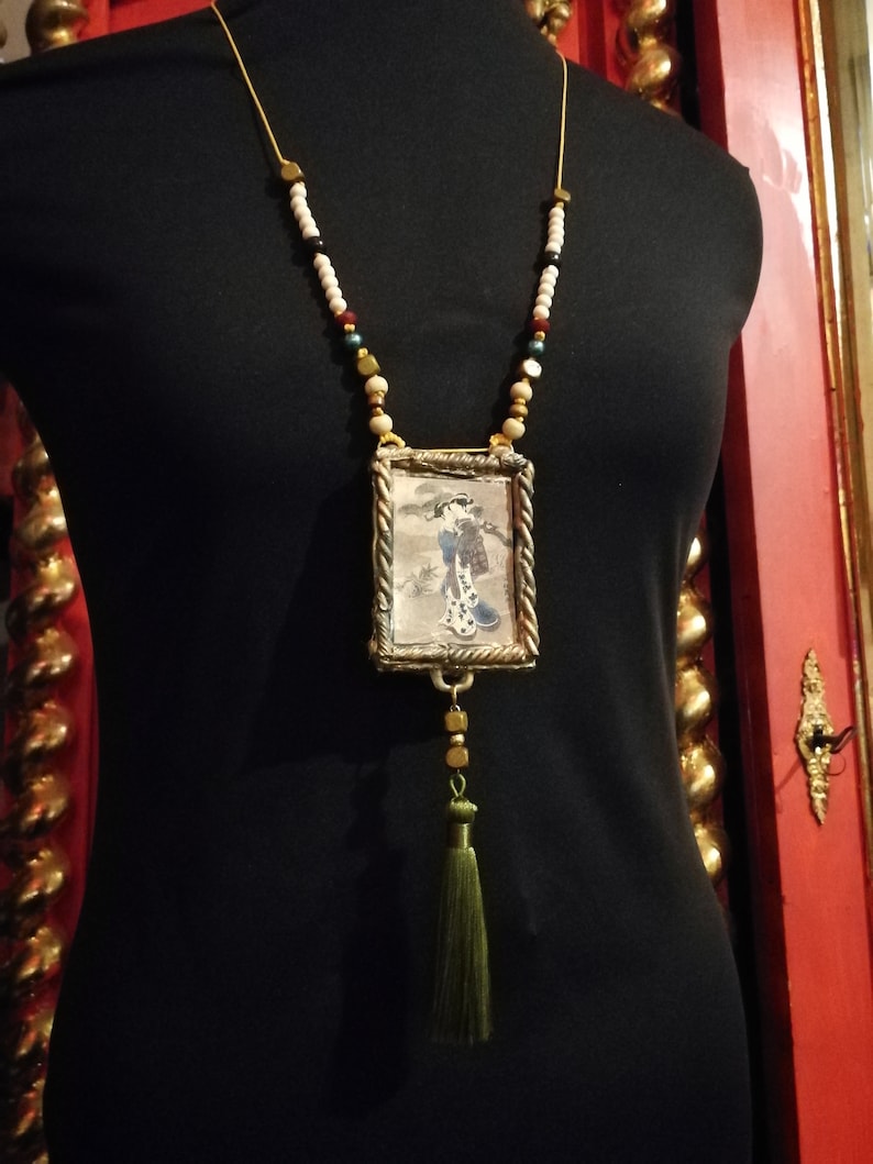 May include: A gold necklace with a small framed picture of a woman in a kimono. The necklace has white, red, and green beads and a green tassel.